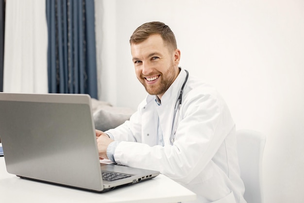 Médico caucásico sentado en el lugar de trabajo y usando una computadora portátil