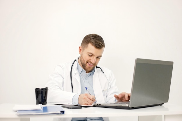 Médico caucásico sentado en el lugar de trabajo y usando una computadora portátil