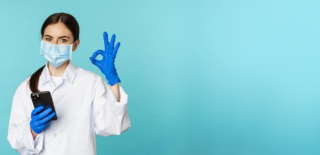 Foto gratuita médico y clínica en línea mujer joven con mascarilla médica que usa un teléfono inteligente para una cita en línea remota del cliente que muestra un signo de aprobación de pie sobre fondo azul