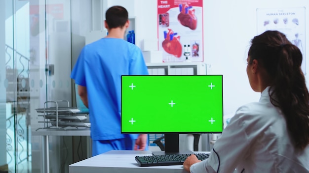 Médico con computadora con clave de croma y asistente con rayos X del paciente en el gabinete del hospital. Escritorio con pantalla verde reemplazable en clínica médica mientras el médico revisa la radiografía del paciente f