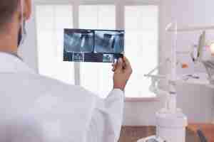 Foto gratuita médico dentista analizando dientes de ortodoncia radiografía médica trabajando en sala de oficina de hospital de estomatología. en el fondo cabient de ortodoncista vacío preparándose para la cirugía de salud de los dientes