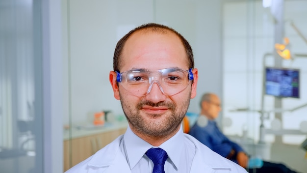 Médico dentista mirando a cámara sonriendo mientras paciente anciano lo espera en segundo plano para la higiene dental. Estomatólogo con gafas de protección de pie frente a la webcam en la clínica estomatológica.