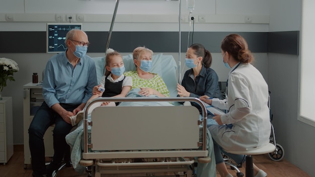 Médico discutiendo sobre el tratamiento con pacientes enfermos en la visita con la familia durante la pandemia del coronavirus. Médico que da consejos médicos a ancianas y familiares con mascarilla en la sala del hospital.