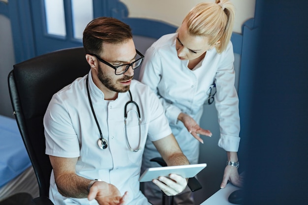 Médico y enfermero masculino examinando registros médicos en el panel táctil y comunicándose en el hospital