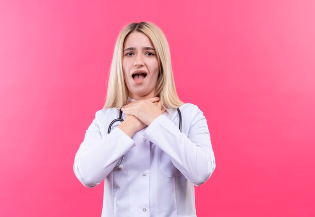 Médico enfermo joven rubia con estetoscopio en bata médica agarró la garganta con ambas manos sobre fondo rosa aislado