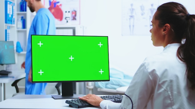 Médico por escrito diagnosticar en computadora con pantalla verde y asistente con uniforme azul en el fondo. Médico en bata blanca trabajando en el monitor con clave cromática en el gabinete de la clínica para revisar al paciente