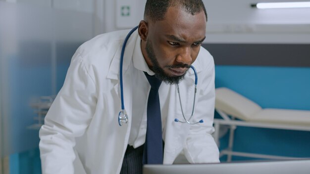 Médico especialista afroamericano sosteniendo un portapapeles analizando el informe de enfermedad comprobando la experiencia médica durante el examen clínico en el consultorio del hospital. Hombre terapeuta que trabaja en el tratamiento de la salud