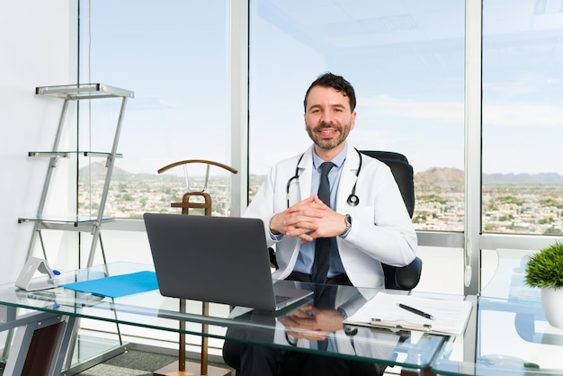 Médico especialista con una gran trayectoria. Guapo doctor sonriendo y haciendo contacto visual mientras está sentado detrás de un escritorio en su oficina con una gran vista de la ciudad