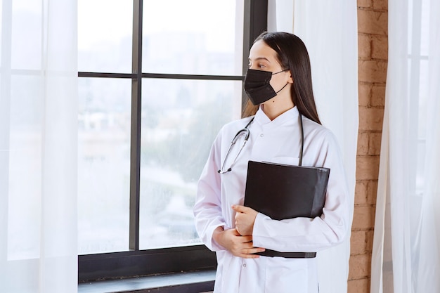 Médico con estetoscopio y máscara negra de pie junto a la ventana y sosteniendo una carpeta de historial negro de los pacientes mientras mira por la ventana.
