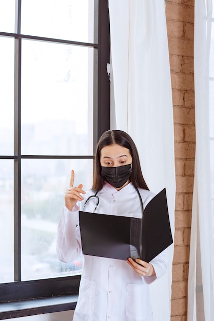 Médico con estetoscopio y máscara negra de pie junto a la ventana y sosteniendo una carpeta negra de la historia de los pacientes, revisándola y se ve aterrorizada.