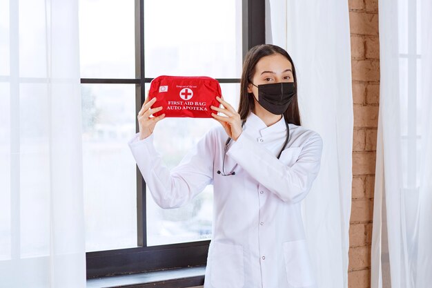 Médico con estetoscopio y máscara negra sosteniendo un botiquín de primeros auxilios rojo.