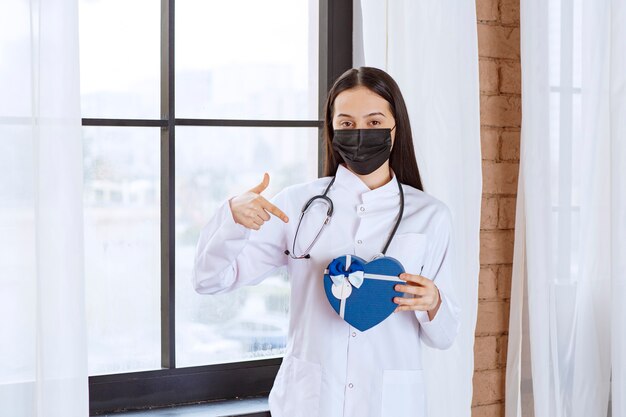 Médico con estetoscopio y máscara negra sosteniendo una caja de regalo con forma de corazón azul.