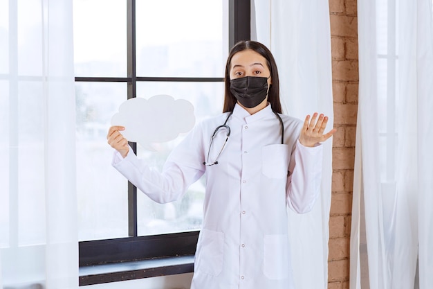 Médico con estetoscopio y máscara negra sosteniendo un panel de ideas con forma de nube blanca y parece confundido.