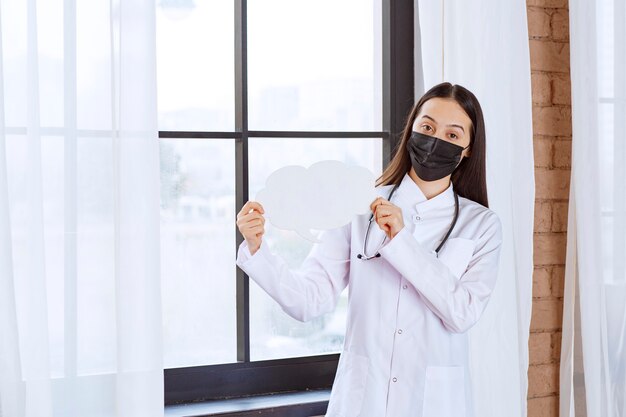 Médico con estetoscopio y máscara negra sosteniendo un panel de ideas con forma de nube blanca.