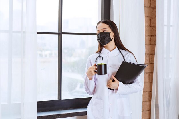 Médico con estetoscopio y máscara negra sosteniendo una taza de bebida negra y una carpeta negra y mirando por la ventana.