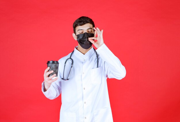 Médico con estetoscopio en máscara negra sosteniendo una taza de café para llevar negra y disfrutando del sabor.
