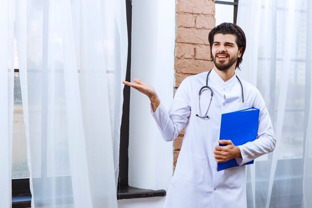 Médico con un estetoscopio sosteniendo una carpeta de informes azul y apuntando a alguien alrededor.