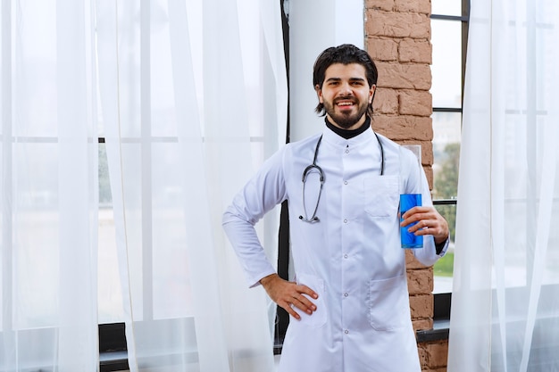 Médico con un estetoscopio sosteniendo un matraz químico con líquido azul en su interior.