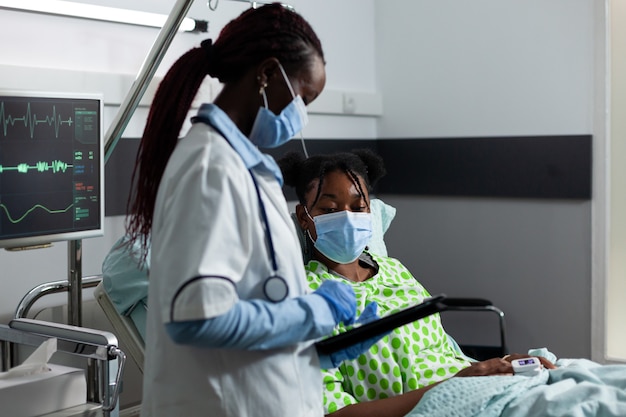 Médico de etnia africana que ayuda al paciente enfermo en la clínica en la sala del hospital. Médico con tecnología y equipos médicos para la atención y el tratamiento de la mujer joven. Persona negra en la cama