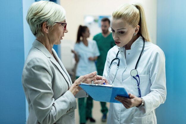 Médico general y mujer madura analizando registros médicos y hablando sobre los resultados de las pruebas en un pasillo de la clínica