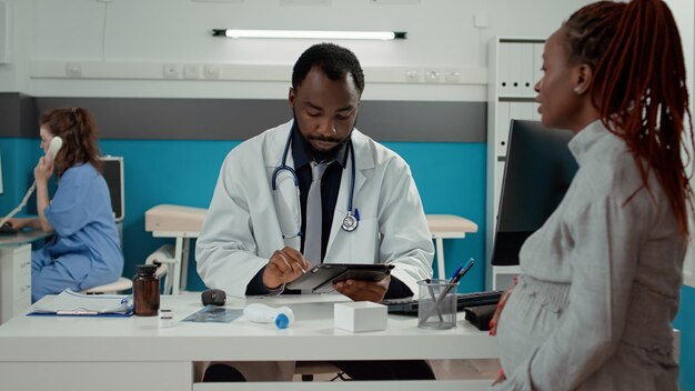 Médico general tomando notas en tableta digital en el chequeo de maternidad con paciente embarazada. Médico masculino conversando sobre el embarazo con la futura madre en el consultorio médico.