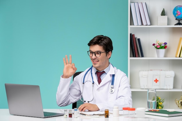 Médico guapo lindo médico en bata de laboratorio que trabaja de forma remota en la computadora que muestra el signo ok