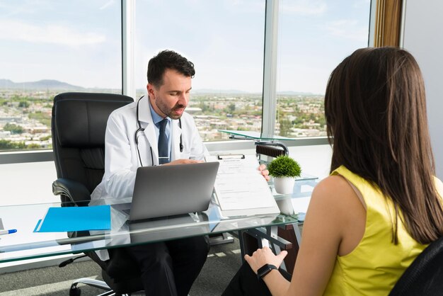 Médico hispano hablando con una paciente en su consultorio y pidiéndole que complete un formulario médico con sus síntomas para encontrarle un tratamiento