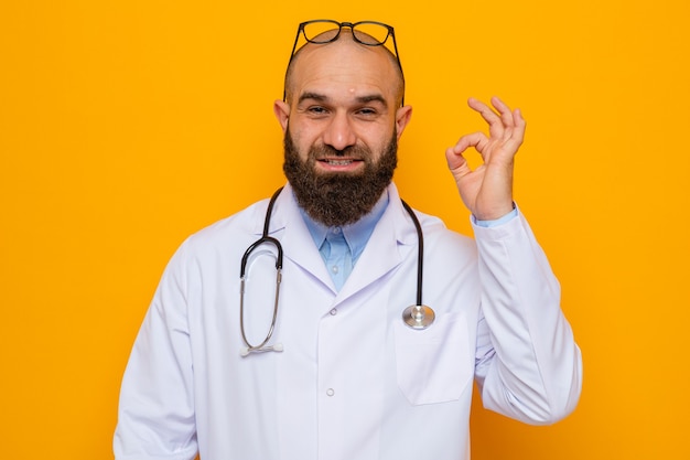 Médico hombre barbudo en bata blanca con estetoscopio alrededor del cuello con gafas en la cabeza mirando a la cámara sonriendo alegremente mostrando signo ok de pie sobre fondo naranja