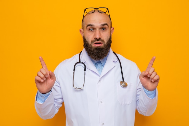 Médico hombre barbudo en bata blanca con estetoscopio alrededor del cuello con gafas en la cabeza mirando a cámara sorprendido apuntando con los dedos índices de pie sobre fondo naranja