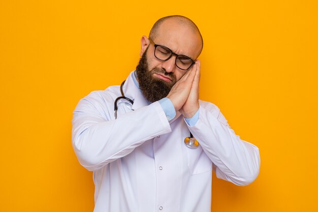 Médico hombre barbudo en bata blanca con estetoscopio alrededor del cuello con gafas cogidos de la mano juntos haciendo gesto de sueño con la cabeza inclinada sobre las palmas quiere dormir de pie sobre fondo naranja