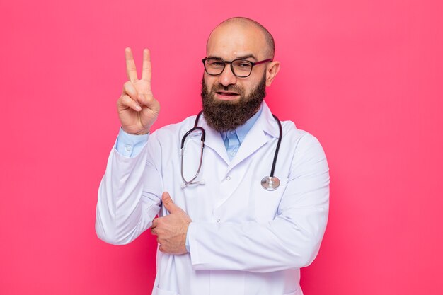 Médico hombre barbudo en bata blanca con estetoscopio alrededor del cuello con gafas mirando a cámara feliz y positivo mostrando signo v de pie sobre fondo rosa