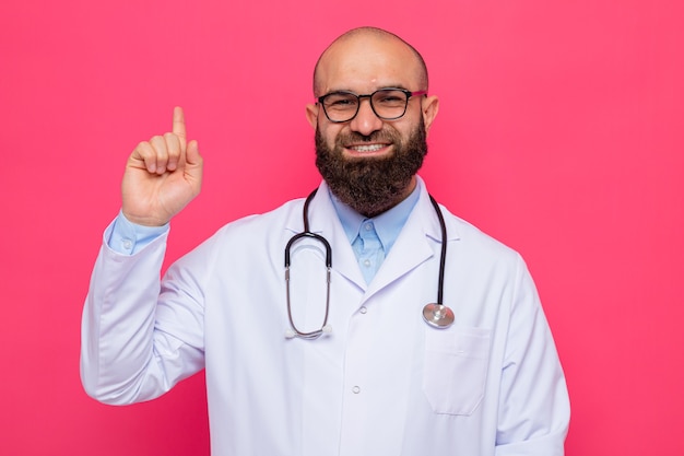 Médico hombre barbudo en bata blanca con estetoscopio alrededor del cuello con gafas mirando a cámara feliz y positivo sonriendo alegremente mostrando el dedo índice de pie sobre fondo rosa