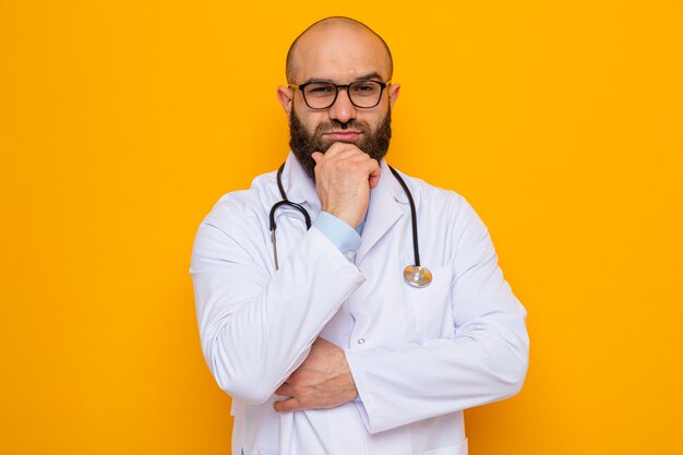 Médico hombre barbudo en bata blanca con estetoscopio alrededor del cuello con gafas mirando a la cámara con la mano en la barbilla pensando sobre fondo naranja