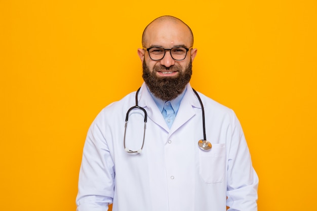 Médico hombre barbudo en bata blanca con estetoscopio alrededor del cuello con gafas mirando a la cámara sonriendo confiado de pie sobre fondo naranja