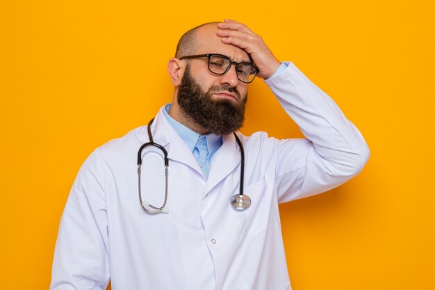 Médico hombre barbudo en bata blanca con estetoscopio alrededor del cuello con gafas mirando confundido y muy ansioso sosteniendo la mano en la cabeza por error de pie sobre fondo naranja