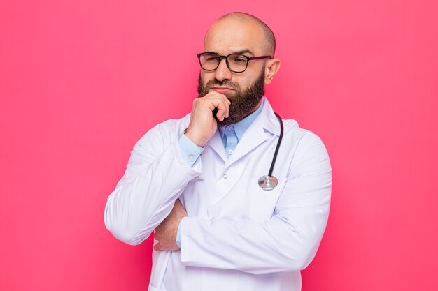 Médico hombre barbudo en bata blanca con estetoscopio alrededor del cuello con gafas mirando a un lado con expresión pensativa con la mano en la barbilla pensando
