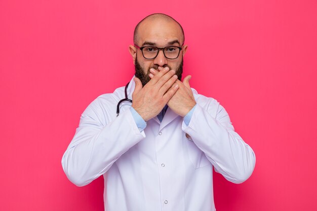 Médico hombre barbudo en bata blanca con estetoscopio alrededor del cuello con gafas mirando sorprendido cubriendo la boca con las manos