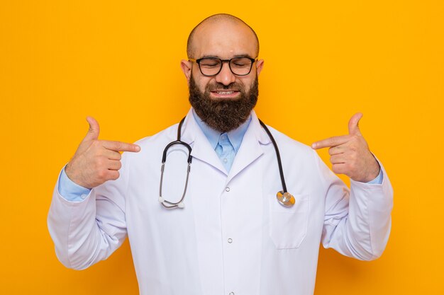 Médico hombre barbudo en bata blanca con estetoscopio alrededor del cuello con gafas sonriendo confiado apuntando a sí mismo