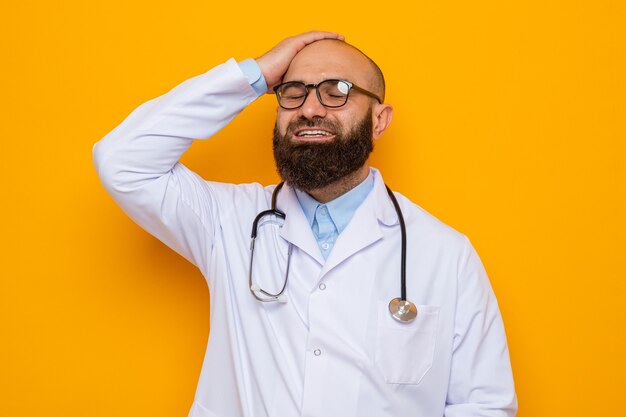 Médico hombre barbudo en bata blanca con estetoscopio alrededor del cuello con gafas sonriendo feliz y emocionado sosteniendo la mano en la cabeza