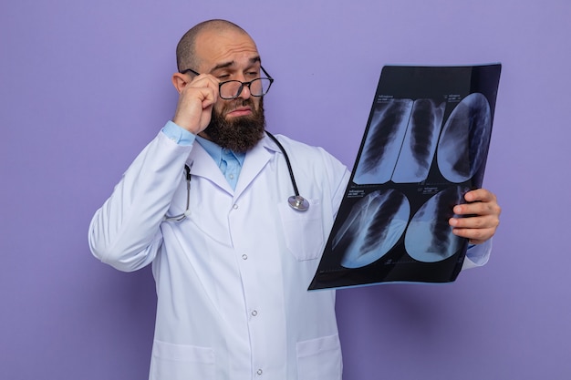 Médico hombre barbudo en bata blanca con estetoscopio alrededor del cuello con gafas sosteniendo rayos x mirándolo de cerca concentrándose de pie sobre fondo púrpura