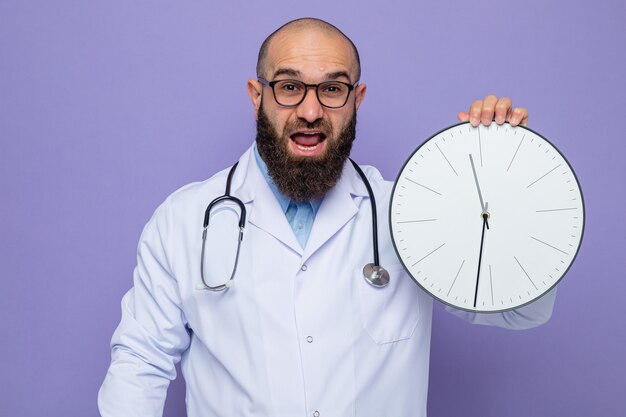 Médico hombre barbudo en bata blanca con estetoscopio alrededor del cuello con gafas sosteniendo reloj mirando a cámara feliz y emocionado de pie sobre fondo púrpura