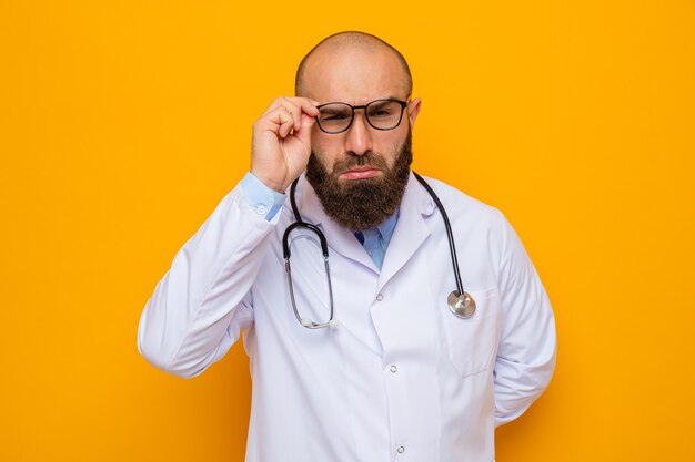 Médico hombre barbudo en bata blanca con estetoscopio alrededor del cuello mirando a la cámara de cerca a través de sus gafas de pie sobre fondo naranja