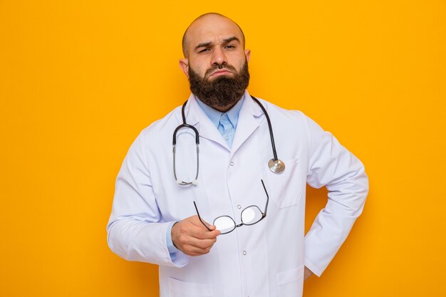 Médico hombre barbudo en bata blanca con estetoscopio alrededor del cuello mirando a la cámara con expresión de confianza sosteniendo gafas de pie sobre fondo naranja