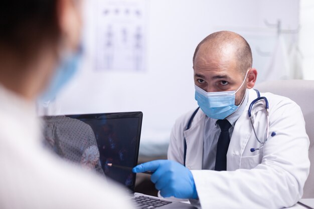 Médico hombre y paciente joven con máscara de protección hablando sobre la evolución del virus sentado en una silla frente a la computadora portátil en la clínica de la oficina del hospital. Trabajador médico que muestra los síntomas apuntando en el escritorio