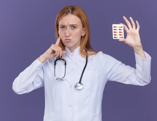 Médico de jengibre joven confundido con bata médica y estetoscopio mirando al frente mostrando el paquete de píldoras médicas al frente tocando la mejilla aislada en la pared púrpura