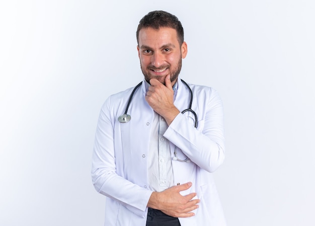 Médico joven en bata blanca y con estetoscopio alrededor del cuello mirando feliz y positivo sonriendo confiado