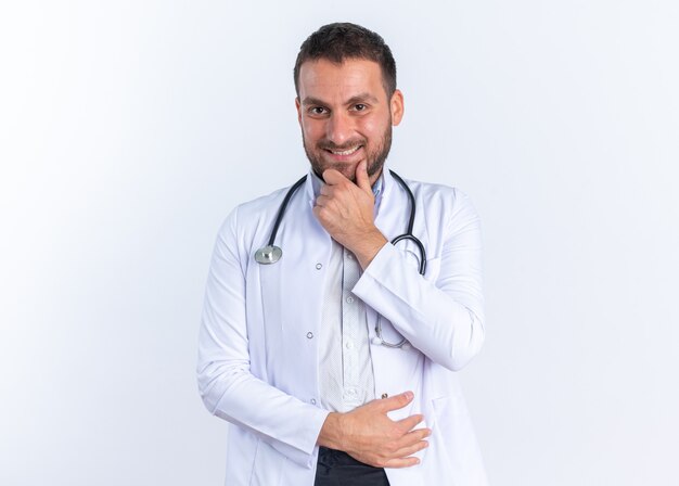 Médico joven en bata blanca y con estetoscopio alrededor del cuello mirando feliz y positivo sonriendo confiado