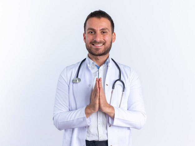 Médico joven en bata blanca y con estetoscopio alrededor del cuello mirando feliz y positivo tomados de la mano juntos como gesto de namaste sonriendo ampliamente