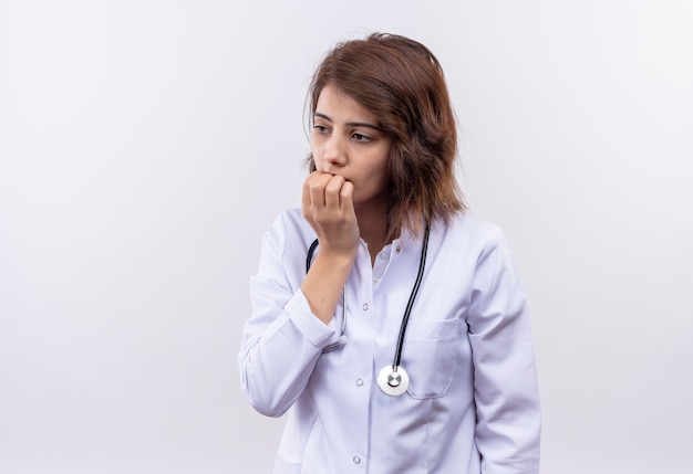 Médico joven en bata blanca con estetoscopio estresado y nervioso morderse las uñas de pie sobre la pared blanca