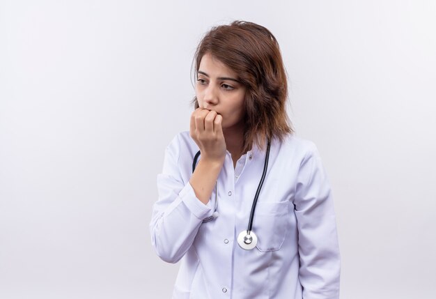 Médico joven en bata blanca con estetoscopio estresado y nervioso morderse las uñas de pie sobre la pared blanca